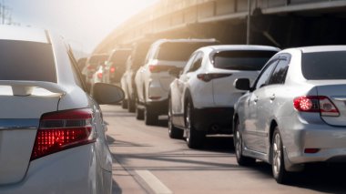 Beyaz arabanın arka tarafında fren lambası açık. Çimento yolunda trafik sıkışık. Birçok araba trafik ışıklarında uzun bir kuyrukta durdu. Arka plan çimento köprüsü üstgeçidi ve parlak gökyüzü.