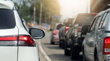 Beyaz arabanın arka tarafında fren lambası açık. Şehirdeki yol trafiği çok sıkışık. Birçok araba trafik ışıklarında uzun bir kuyrukta durdu. Arkaplan ağaçları ve beton köprü üstgeçidi.