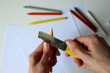 Renkli kalemleri büro bıçağıyla açarken çekilmiş bir fotoğraf. İnsan eli. Sarı kalem. Metal bıçak. Arka planda beyaz kağıtlar ve renkli kalemler var. Yaratıcılık, çizim, sanat, sanatçı.