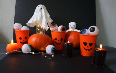 Halloween decor. Set of different handmade elements on a black background. Orange jack o lantern cups filled with eyes. Ghost from a napkin. Pumpkins. Black candles sprinkled with red drops. Garland. clipart