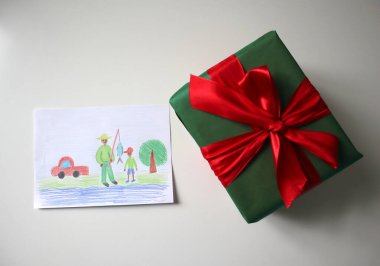 Bir çocuğun çizdiği kartpostalın Babalar Günü fotoğrafı. Kırmızı fiyonklu yeşil bir hediye. Beyaz arka plan. Kartpostalda baba oğul balık tutarken görülüyor. Afrika görünüşü. Renkli kalemlerle çizim.