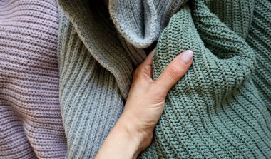 Örgü ören bir kadın eli. Arka planda mor, gri, turkuaz renkli örgü kumaşlar var. Kaotik bir yer. El, turkuaz bir bez tutuyor. Başparmak vernikle kaplıdır. Manikür.