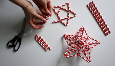 El yapımı. Kadın eli iplikle iğneyi tutuyor. Kokteyller için kağıt tüplerden Noel yıldızları yaratma sürecinde. Kırmızı, beyaz çizgili tüpler. Makas, beyaz iplik. Beyaz arka plan. Fotoğraf.