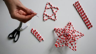 El yapımı. Kadın eli iplikle iğneyi tutuyor. Kokteyller için kağıt tüplerden Noel yıldızları yaratma sürecinde. Kırmızı beyaz çizgili tüpler. Makas, beyaz iplik. Beyaz arka plan. Fotoğraf.