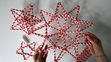 Dişi eller yıldızı tutar. Beyaz arka plan, diğer yıldızlar, iğne, beyaz iplik. Bütün yıldızların farklı şekilleri vardır. El yapımı dekor. Kırmızı, beyaz şeritli tüplerden yapılmış. Ellerinde gümüş aksesuarlar. Fotoğraf.