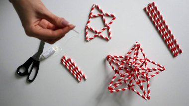 Kadın eli iplikle iğneyi tutuyor. Kokteyller için kağıt tüplerden yıldız yapma süreci. Kırmızı beyaz çizgili tüpler. Makas, beyaz iplik. Beyaz arka plan. Fotoğraf. Arkaplana odaklan.