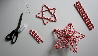 Kokteyller için pipetten yapılmış yıldızlar. Kırmızı ve beyaz çizgili. İğne ve beyaz iplik, makas, farklı uzunlukta kokteyl tüpleri. Beyaz arka planda. El yapımı. Dekorasyon için fikirler. Noel zamanı. ÇEVİR