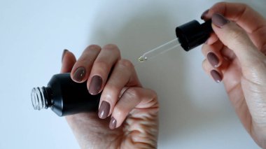 Bir kadının eli küçük siyah bir şişe tutuyor. Diğer yandan pipetle parmağına yağ damlatıyor. Tırnaklar manikürle kaplı. Açık kahverengi renk. Beyaz arka plan. Beyaz eller. Makro fotoğraf.