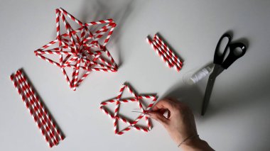 Kadın eli iplikle iğneyi tutuyor. Kokteyller için kağıt tüplerden yıldız yapma süreci. Kırmızı beyaz çizgili tüpler. Makas, beyaz iplik. Beyaz arka plan. Fotoğraf. Arkaplana odaklan.