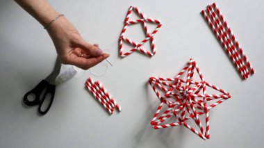 El yapımı. Kadın eli iplikle iğneyi tutuyor. Kokteyller için kağıt tüplerden Noel yıldızları yaratma sürecinde. Kırmızı beyaz çizgili tüpler. Makas, beyaz iplik. Beyaz arka plan. Fotoğraf.