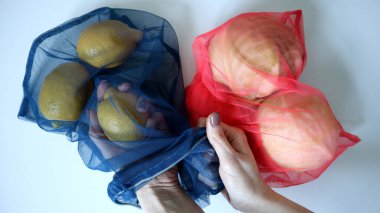 Meyve ve sebzelerin ekolojik ağ torbalarında fotoğrafı. Beyaz arka plan. Mavi çantada üç limon. Limonu çıkaran kadın elleri. Pembe çantada iki beyaz lahana. Ürünler, alışveriş, sağlıklı beslenme..