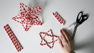 Kadın eli iplikle iğneyi tutuyor. Kokteyller için kağıt tüplerden yıldız yapma süreci. Kırmızı beyaz çizgili tüpler. Makas, beyaz iplik. Beyaz arka plan. Fotoğraf. Arkaplana odaklan.