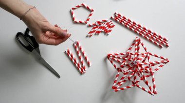 El yapımı. Kadın eli iplikle iğneyi tutuyor. Kokteyller için kağıt tüplerden Noel yıldızları yaratma sürecinde. Kırmızı beyaz çizgili tüpler. Makas, beyaz iplik. Beyaz arka plan. Fotoğraf.