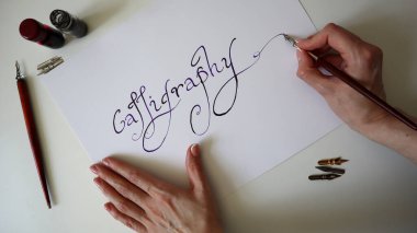 Kaligrafi süreci. Kaligrafi kelimesini yazan kadın el yazısı. Kalemler, mürekkep yakınlarda. Yazı tipi mor ve siyah renk. Beyaz kadın elinde kalem, diğer elinde kağıt var. Beyaz arka plan. Fotoğraf..