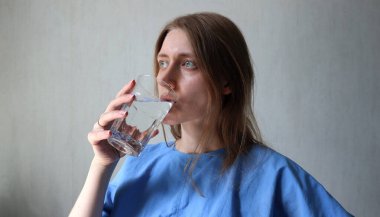 Su içen kadın. Kafkasyalı, sarı saçlı. Mavi gözlü ve gömlekli. Temiz bir bardak su tutuyorum. Makyaj yok, doğal görünüş. Fransız manikürü. Sola bak. Açık gri arkaplan. Fotoğraf..