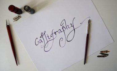 Kaligrafi kelimesi beyaz kağıda el yazısıyla yazılmış. Etrafta dolma kalem ve mürekkep var. Yazı tipi yuvarlak, uzun serifler. Mordan siyaha doğru bir eğim var. Beyaz arka plan. Fotoğraf..