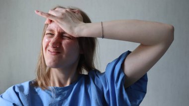 Baş ağrısı, endişe, endişe. Acımasız bir ifadeyle başını eliyle tutan bir kadın. Kafkasyalı, sarı saçlı. Üzücü bir ifade. Elin alnında. Mavi bir gömlek giyiyor. Açık gri arkaplan. Fotoğraf..