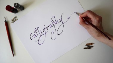 Kaligrafi süreci. Kadın el yazısı Kaligrafi. El yazısı yazı tipi. Kalemler, mürekkep yakınlarda duruyor. Yazı tipi siyahtan mora doğru eğimli. Beyaz kadın kalem tutuyor. Beyaz arka plan. Fotoğraf..