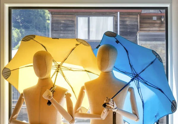 Two jointed mannequins inside a room, holding a yellow and a blue umbrella up, with a clear window and bright interior backdrop.