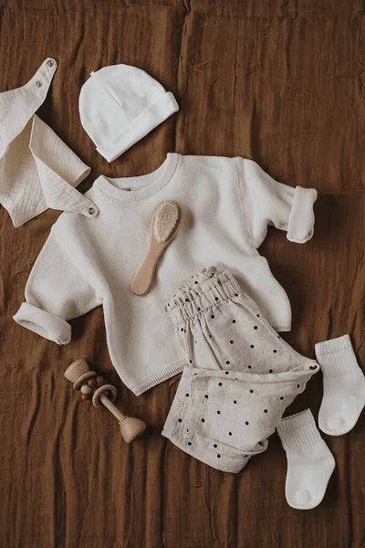 Cute hygge pastel clothes, accessories for newborn baby. Muslin bib, sweater, joggers, socks, brush, hat on brown linens. Aesthetic luxury baby fashion store, shopping concept. Flatlay, top view