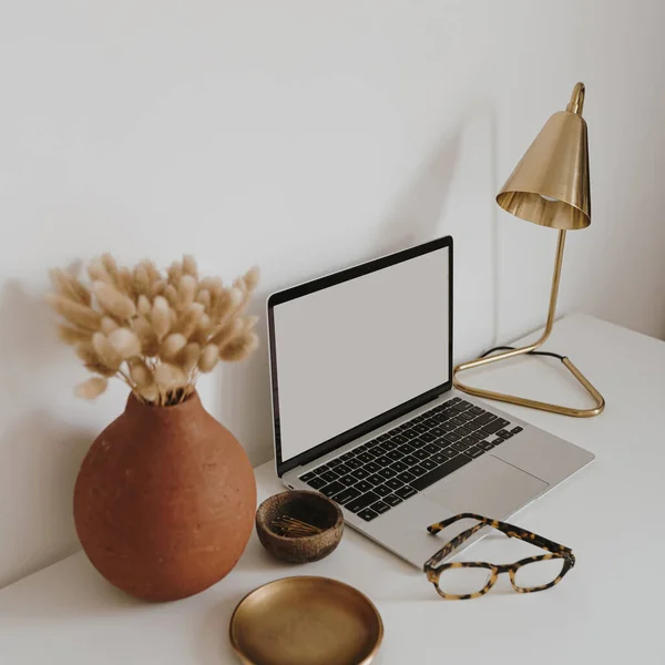 Aesthetic minimalist home office workspace desk. Laptop computer with blank copy space display screen, glasses, dried grass bouquet, lamp, golden plate. Chic lady boss business branding concept