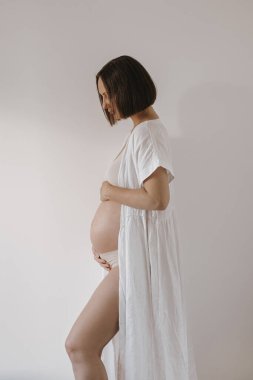 Genç hamile kadın beyaz duvarın yanında kalıyor. Hamilelik, annelik, annelik kavramı. Koca göbekli mutlu hamile kadın.