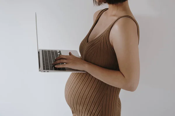 身穿棕色衣服 手持笔记本电脑的年轻漂亮的孕妇靠在白墙边 — 图库照片