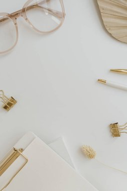 Frame of elegant neutral golden stylish home office desk with glasses, fan leaf stem, clipboard, accessories on white background. Aesthetic flat lay, top view workspace with blank copy space