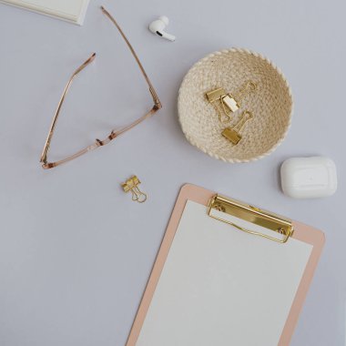 Flatlay of clipboard with blank copy space paper sheet. Glasses, wireless headphones, clips on pastel background. Home office desk workspace. Aesthetic business, work template. Flat lay, top view
