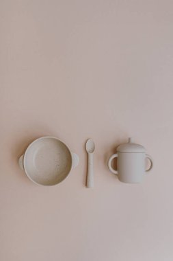 Sevimli bebek sofra takımı düzleştirilmiş. Tabak, bardak, tarafsız arka planda kaşık. Bebek ilk yemek aksesuarlarını servis ediyor. Düz yatış, üst görünüm beslenme ve beslenme konsepti