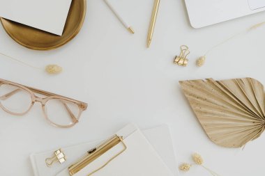 Frame of elegant neutral golden stylish home office desk with glasses, fan leaf stem, clipboard, accessories on white background. Aesthetic flat lay, top view workspace with blank copy space