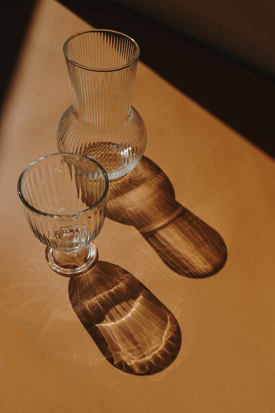 Эстетическая богемная элегантная композиция с винтажной стеклянной вазой и бокалом шампанского с размытыми силуэтами солнечного света на теплом оранжевом фоне