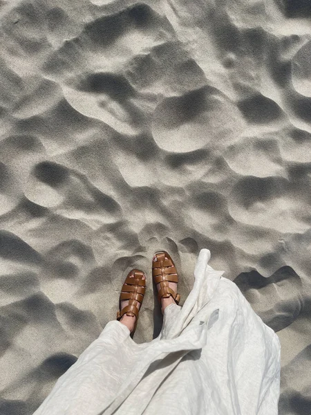 stock image Women feet on desert beach sand. Aesthetic summer vacation background. Woman in white dress and leather sandals on the dune sand. Top view