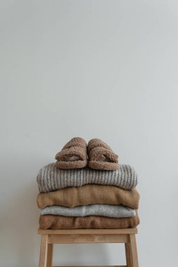 Beyaz duvarın üzerindeki tahta taburede sıcak, nötr bej giysiler yığını. Yumuşak terlikler, yün kazaklar ve kazaklar. Minimalist sonbahar, sonbahar moda geçmişi
