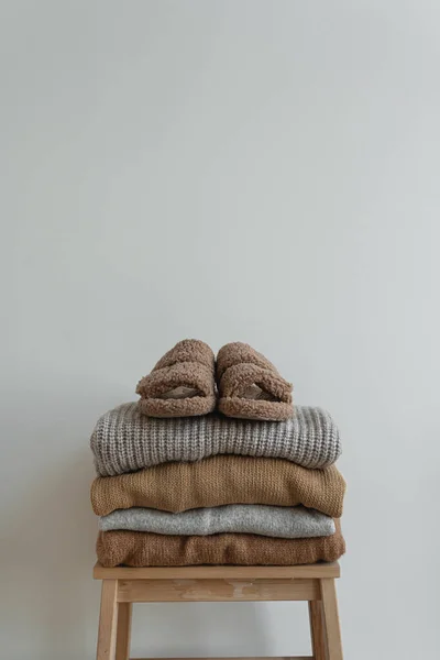 Stack of warm neutral beige clothes on wooden stool over white wall. Fluffy slippers, wool pullovers and sweaters. Minimalist autumn, fall fashion background