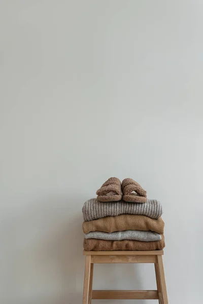 Stack of warm neutral beige clothes on wooden stool over white wall. Fluffy slippers, woolen pullovers and sweaters
