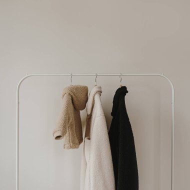 Sonbahar kıyafetleri beyaz duvardan sarkıyor. Sıcak bebek ve yetişkin ceketleri. Kıyafetleri tarafsız renklerde olan estetik bir gardırop.