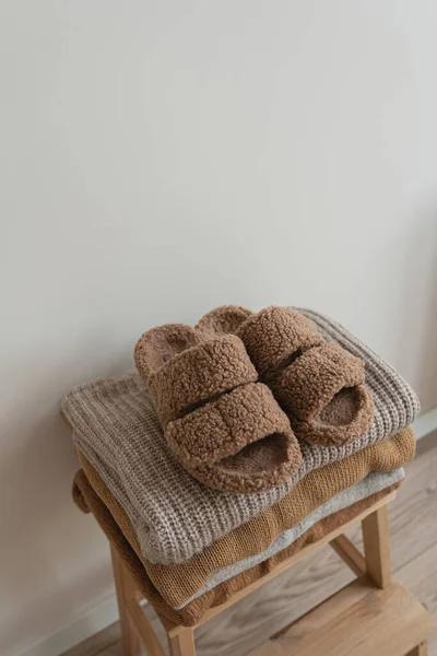 Stack of warm neutral beige clothes on wooden stool over white wall. Fluffy slippers, woolen pullovers and sweaters