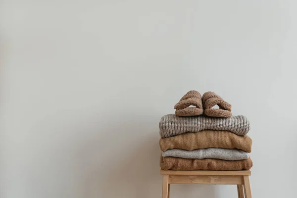 Stack of warm neutral beige clothes on wooden stool over white wall. Fluffy slippers, wool pullovers and sweaters. Minimalist autumn, fall fashion background