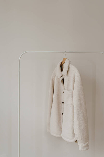 Белая тёплая шерстяная осенняя куртка на вешалке над белой стеной. Минималистский гардероб