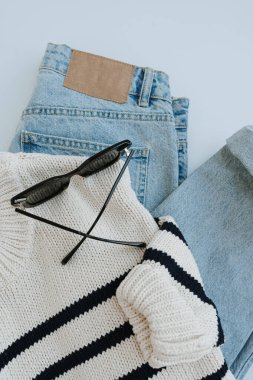 Kadın kıyafetleri ve aksesuarlarıyla minimalist estetik günlük moda kompozisyonu. Çizgili kazak, kot pantolon, güneş gözlüğü. Düz yatış, üst görünüm.