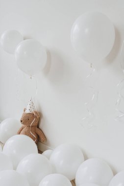Parti şapkalı oyuncak ayı beyaz arka planda balonların yanında oturuyor. Çocukların doğum günü partisi. Bebeğin birinci yıl dönümü