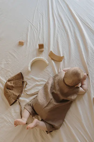 新生児がベッドの上に寝そべっている様子 かわいい居心地の良いリネンのボディスーツ 木製のおもちゃ 小さな赤ちゃんのファッションコンセプト — ストック写真