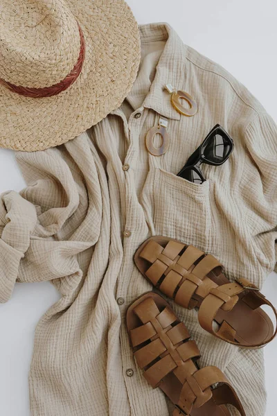 Aesthetic summer fashion composition with neutral beige female clothes and accessories. Muslin tee blouse, straw hat, leather sandals, sunglasses, earrings. Flat lay, top view.