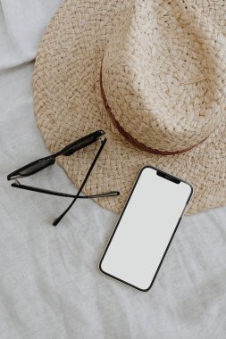 Estetik şık internet mağazası, online alışveriş, moda blogu, sosyal medya markalaşma kompozisyonu. Nötr bej kumaş, hasır şapka ve güneş gözlüklü boş kopya alan ekranlı cep telefonu