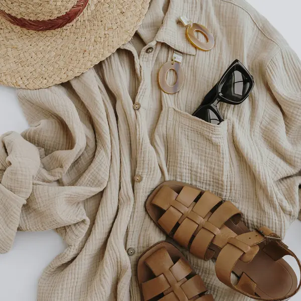 Aesthetic summer fashion composition with neutral beige female clothes and accessories. Muslin tee blouse, straw hat, leather sandals, sunglasses, earrings. Flat lay, top view.