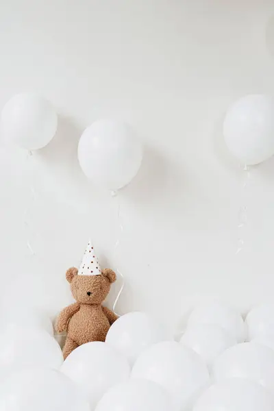 Teddy bear in party hat sitting near balloons over white background