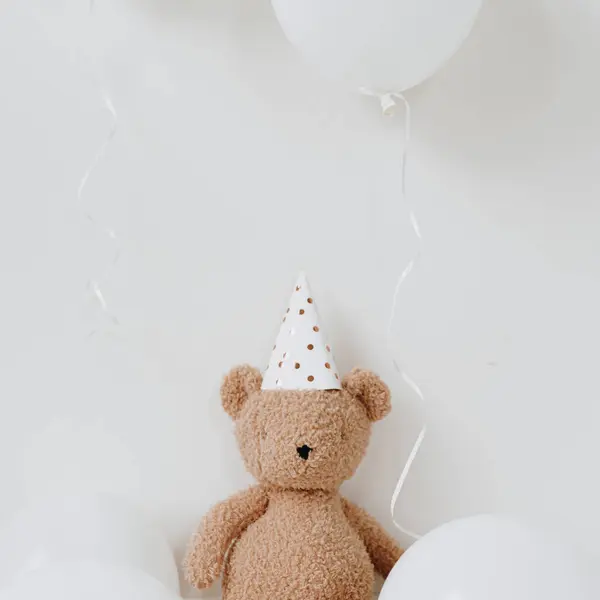 Cute Teddy Bear toy in party hat sitting near balloons over white background. Children\'s birthday party celebration. Baby\'s first year anniversary. Balloon with blank copy space