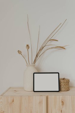 Boş ekranlı tablet tableti, kuru otlu kil tabut ve beyaz duvarda haşhaş sapı. Sosyal medya için estetik nötr bej şablonu, model için kopyalama alanı olan mobil web sitesi