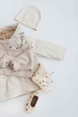 Yağmurlu havalar için bej renkli çocukların sıcak giysileri. Çocuk süveteri, yağmurluk, lastik çizmeler, beyaz arka planda şapka. Düz yatıyordu, İskandinav Bebek Moda Kolajı